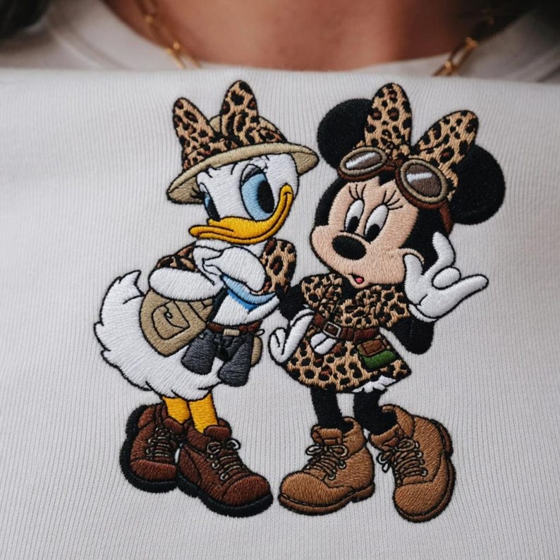 Pu&ograve; includere: Design ricamato con Paperino e Minnie Mouse in tenute safari. Paperino indossa un cappello safari, un gilet e stivali. Minnie Mouse indossa un vestito con stampa leopardata, occhiali e stivali. Il design &egrave; su sfondo bianco.