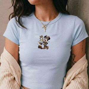 Pu&ograve; includere: T-shirt azzurra con una grafica di Minnie e Paperina in abiti coordinati con stampa leopardata. I personaggi dei cartoni animati si abbracciano. La maglietta ha maniche corte e girocollo.