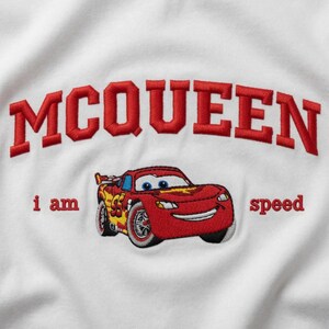 May include: White t-shirt featuring an embroidered design of Lightning McQueen from the Cars movie. The design includes the word "MCQUEEN" in red, arched lettering above the car, and the phrase "i am speed".