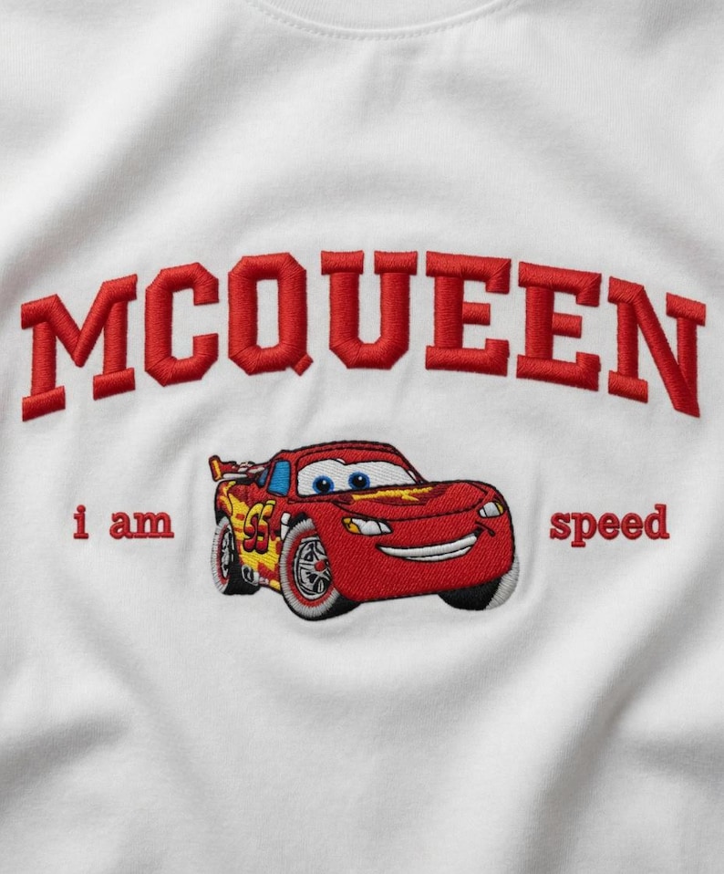 May include: White t-shirt featuring an embroidered design of Lightning McQueen from the Cars movie. The design includes the word "MCQUEEN" in red, arched lettering above the car, and the phrase "i am speed".