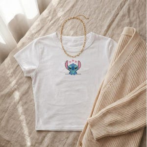 Puede incluir: Top corto blanco con un dise&ntilde;o de dibujos animados de Stitch y corazones rosas. Una cadena dorada y una rebeca beige tambi&eacute;n est&aacute;n en la imagen. La parte superior est&aacute; hecha de un material suave, adecuado para uso casual.