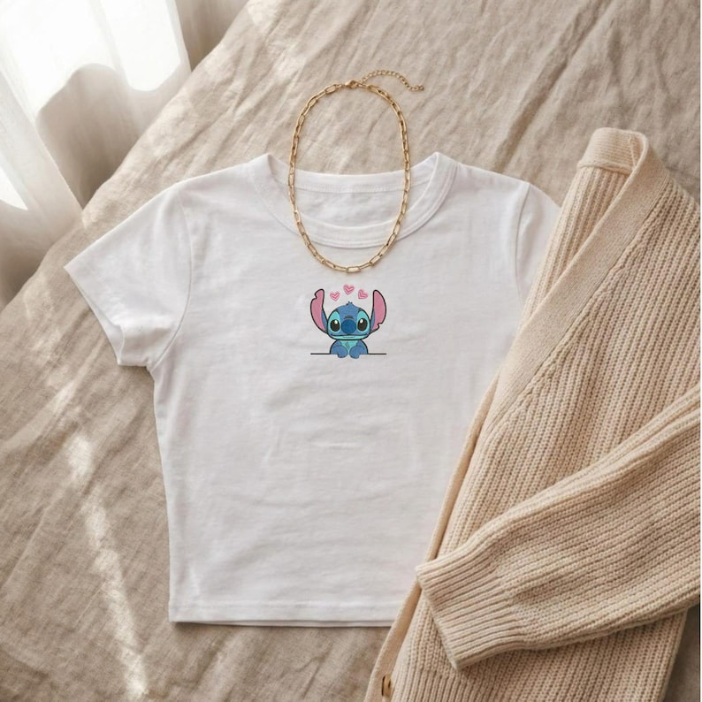 Puede incluir: Top corto blanco con un dise&ntilde;o de dibujos animados de Stitch y corazones rosas. Una cadena dorada y una rebeca beige tambi&eacute;n est&aacute;n en la imagen. La parte superior est&aacute; hecha de un material suave, adecuado para uso casual.