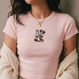 Pu&ograve; includere: Una maglietta rosa chiaro con una grafica di Minnie Mouse e Paperina. I personaggi dei cartoni animati indossano abiti con stampa leopardata abbinati. La maglietta ha maniche corte e girocollo.