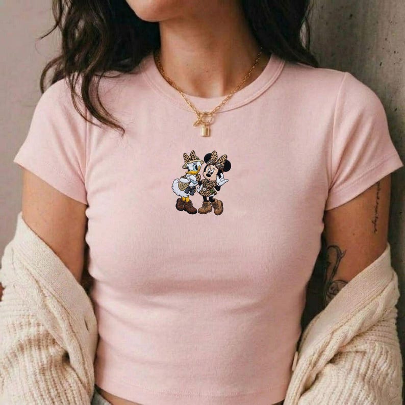 Pu&ograve; includere: Una maglietta rosa chiaro con una grafica di Minnie Mouse e Paperina. I personaggi dei cartoni animati indossano abiti con stampa leopardata abbinati. La maglietta ha maniche corte e girocollo.