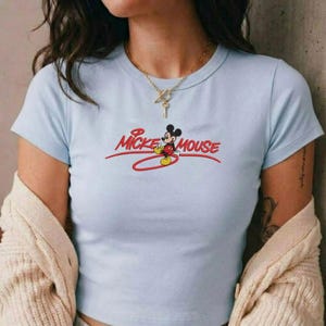 Op de afbeelding: Lichtblauw crop top met de tekst "Mickey Mouse" in rode letters en een cartoonafbeelding van Mickey Mouse. De top heeft korte mouwen en een ronde halslijn.