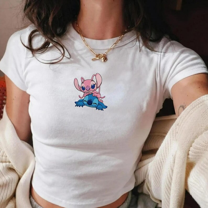 May include: White t-shirt with a cartoon graphic of two characters, one pink and one blue. The pink character is on top of the blue character. The t-shirt has short sleeves and a crew neck.
