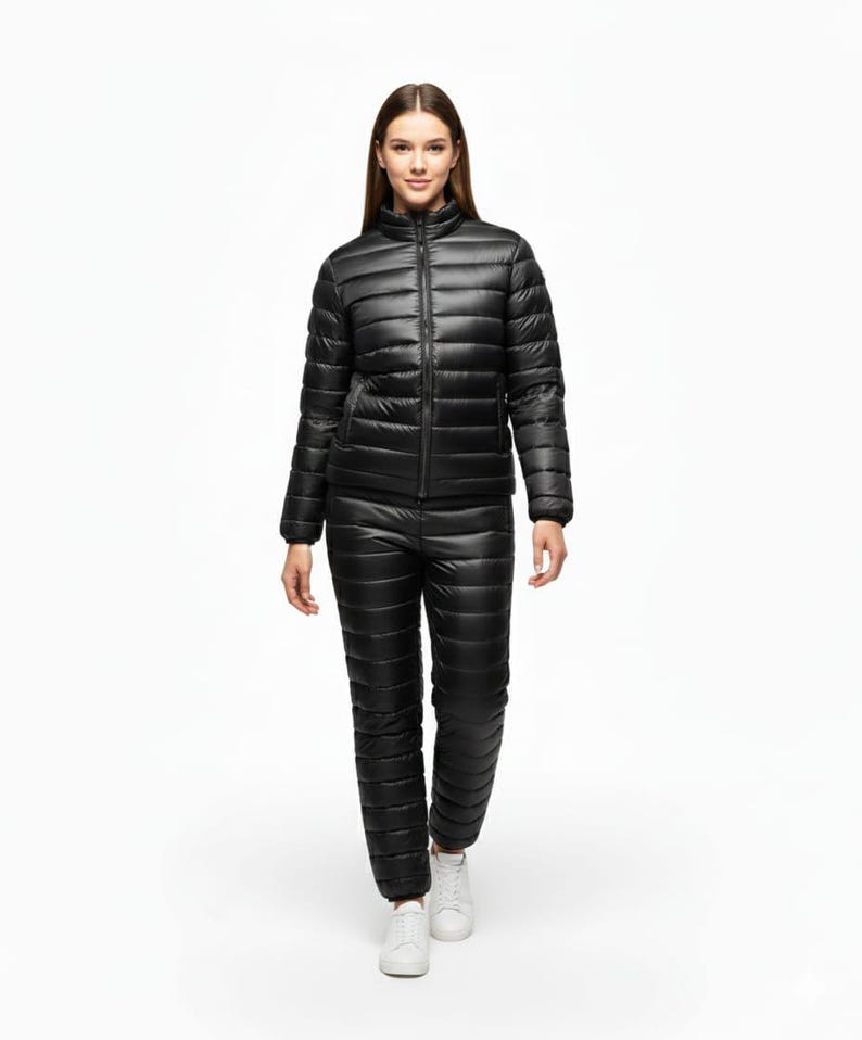 May include: A black quilted puffer jacket and matching pants set. The jacket has a high collar and a full-length zipper. The pants are also quilted and have a straight leg. The person is wearing white sneakers.