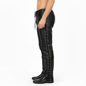 Könnte beinhalten: Schwarze Lederhose mit Schnürdetails an der Seite und vorne. Die Hose hat eine figurbetonte Silhouette und goldfarbene Ösen. Die Hose wird mit schwarzen Stiefeln gezeigt.