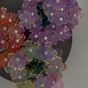 Puede incluir: Una colección de coloridos adornos florales translúcidos con ribetes dorados y centros perlados. Las flores están dispuestas en un patrón circular, con tonos de morado, naranja, rosa y verde. El arreglo se encuentra sobre una superficie gris oscuro.