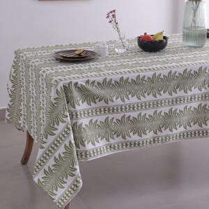 May include: A rectangular white tablecloth with a repeating green leaf pattern. The table is set with a dark plate, gold utensils, and a small bowl of fruit. A vase of flowers and a decorative wall hanging are also visible.
