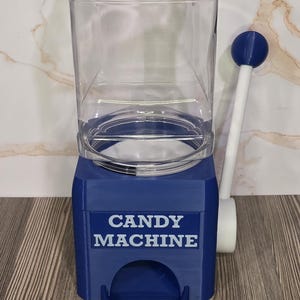 May include: A blue candy machine with a clear plastic container on top and a white lever with a blue ball handle on the side. The front of the blue base features the text CANDY MACHINE in white letters above a circular dispensing opening.