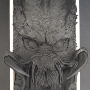 May include: A detailed, black sculpture of a Predator's head, featuring intricate textures and sharp mandibles. The sculpture is set within a white rectangular frame, creating a striking contrast against the dark background.