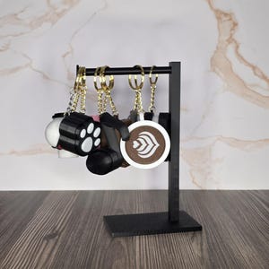 May include: A black metal display stand holding several keychains. The keychains feature various designs including a black and white paw print, a black camera-shaped charm, and a round charm depicting a coffee cup with latte art.