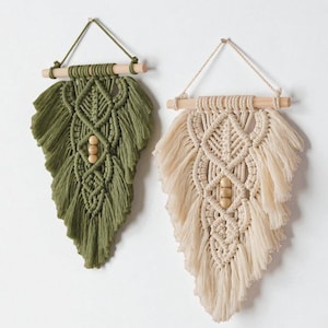 May include: Two macrame wall hangings, one olive green and one cream, each shaped like a feather. They are suspended from wooden dowels with a triangle of string. The designs feature intricate knotting and wooden bead accents.