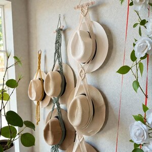 May include: A selection of straw hats displayed on a wall, suspended by macrame hangers in cream and olive green. The hats vary in style and colour. The wall is a neutral tone. The scene is well-lit, creating a bright and airy feel.