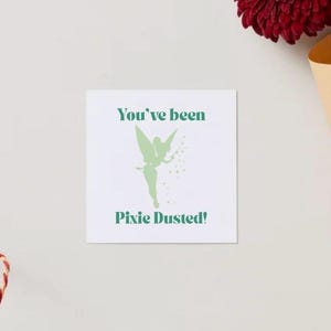 Op de afbeelding: Een vierkante witte kaart met de tekst "You've been Pixie Dusted!" in het groen, en een groen silhouet van een fee. Een rood-wit touwtje en een bordeauxrode bloem in een beige papieren kegel zijn ook zichtbaar.