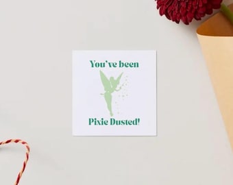 May include: A square white card with the text "You've been Pixie Dusted!" in green, along with a green silhouette of a fairy. A red and white string and a burgundy flower in a tan paper cone are also visible.