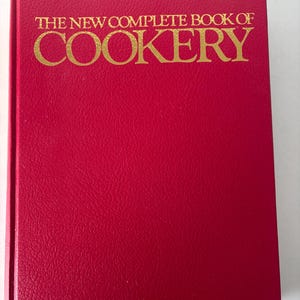 May include: A hardback book featuring a textured red cover with gold embossed text on the front reading THE NEW COMPLETE BOOK OF COOKERY. The book is rectangular in shape, with a vertical spine on the left and a uniform, solid red front surface.