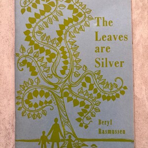 Pode incluir: Capa de livro vintage intitulada "The Leaves are Silver" de Beryl Rasmussen. A capa apresenta uma ilustração verde de uma árvore com folhas e uma silhueta familiar em um fundo azul claro. O texto é verde.