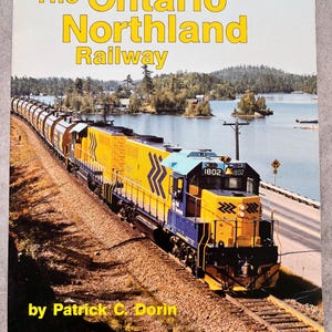 May include: The cover of a book, "The Ontario Northland Railway", shows a yellow and blue train on a track. The background features a lake and trees. The author's name, Patrick C. Dorin, is printed at the bottom of the cover.