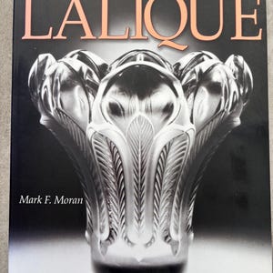 May include: A book cover featuring the word "LALIQUE" in large, peach-colored letters. Below the title is a detailed, grayscale image of a decorative glass vase. The book is titled "Warman's Lalique Identification and Price Guide" by Mark F. Moran.