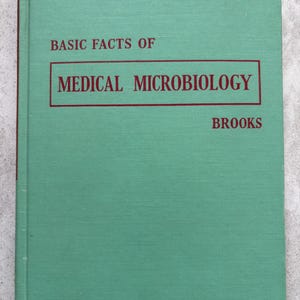 May include: A hardcover book titled "Basic Facts of Medical Microbiology" by Brooks. The book has a teal green cover with the title in a red rectangular box. The text is in a serif font.