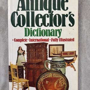 Pode incluir: Um livro vintage intitulado "Antique Collector's Dictionary" com texto verde e branco. A capa apresenta ilustrações de móveis antigos, uma pintura de um navio e um prato decorativo. Publicado pela Info Books.