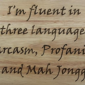 May include: A light-colored wooden board featuring dark, laser-engraved text that reads: I'm fluent in three languages Sarcasm, Profanity, and Mah Jongg. The wood grain is visible throughout the surface, providing a natural texture to the background.