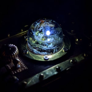 May include: A steampunk-style box with a glass dome containing a swirling, white, light-emitting object. The box is made of dark wood and brass with intricate details.