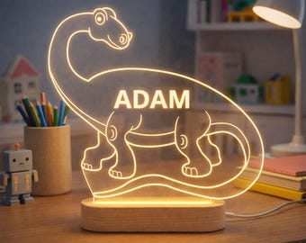 Puede incluir: Una lámpara de noche con forma de dinosaurio brillante con el nombre "ADAM" en blanco. La luz está hecha de acrílico transparente y se asienta sobre una base de madera. El diseño muestra un Braquiosaurio. La luz está sobre un escritorio de madera.