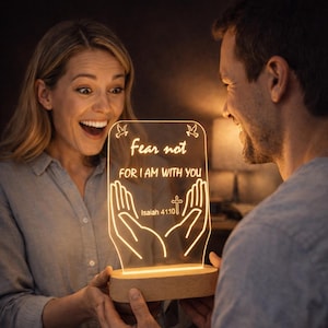 Può includere: Una luce notturna a LED luminosa con le parole "Fear not FOR I AM WITH YOU" e un disegno a croce e mani. La luce è posta su una base di legno ed è tenuta da due persone.