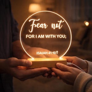 Può includere: Un'insegna rotonda e illuminata con le parole "Fear not, FOR I AM WITH YOU;" e "ISAIAH 41:10†" in scrittura bianca. L'insegna è tenuta da due mani e poggia su una base di legno. Un cavo USB bianco è collegato alla base.