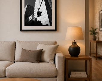 May include: A monochrome print of city buildings hangs above a beige sofa with brown cushions. A wooden side table with a lamp and books sits beside the sofa. A round coffee table and a rug are in the foreground, creating a cozy living space.