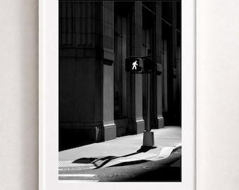 May include: Black and white framed photograph of a city street scene. A pedestrian crossing signal shows a walking figure. The image captures architectural details, shadows, and a traffic cone.