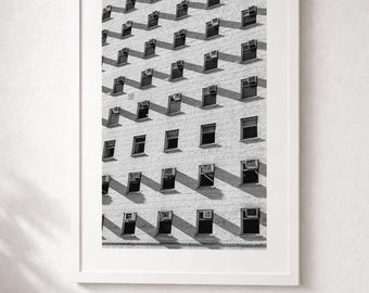 May include: A black and white framed wall art print featuring a repeating architectural pattern of a brick building facade. The image displays multiple rows of rectangular windows, each equipped with a window air conditioning unit.