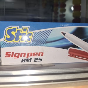 Puede incluir: Una caja de rotuladores Stic Signpen BM 25. La caja es azul con texto blanco y amarillo. En la caja se muestra un rotulador blanco con tapa roja. El rotulador tiene una punta negra.