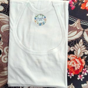 May include: A folded white baby vest with a round neckline and a small, circular sticker that reads "Thank you" in script. The sticker is surrounded by a floral design. The vest is on a patterned fabric.