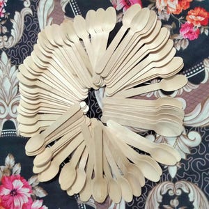 Puede incluir: Un conjunto de cucharas de madera desechables dispuestas en un patrón circular. Las cucharas son de color marrón claro y parecen estar hechas de una sola pieza de madera. El fondo es una tela oscura con patrones florales y ornamentados.