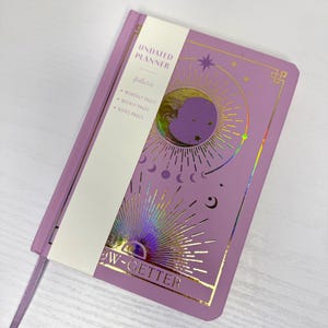 May include: A lilac undated planner with gold foil celestial designs, including a moon and stars. The planner features monthly, weekly, and notes pages. A white band across the front reads "UNDATED PLANNER" and "NOW-GETTER". A lilac ribbon bookmark is included.