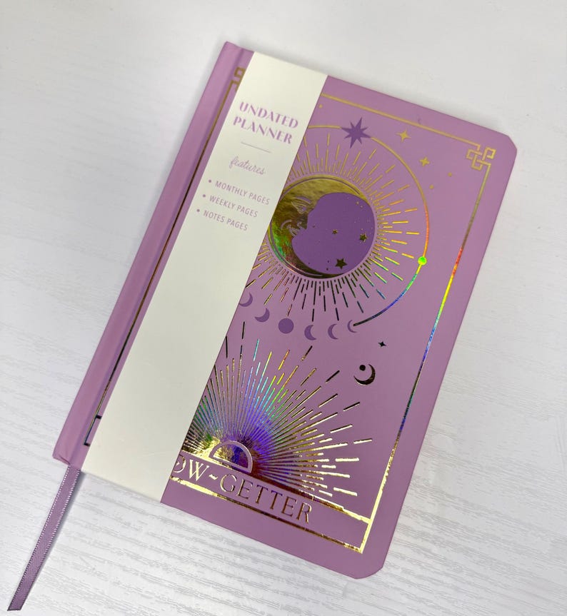 May include: A lilac undated planner with gold foil celestial designs, including a moon and stars. The planner features monthly, weekly, and notes pages. A white band across the front reads "UNDATED PLANNER" and "NOW-GETTER". A lilac ribbon bookmark is included.