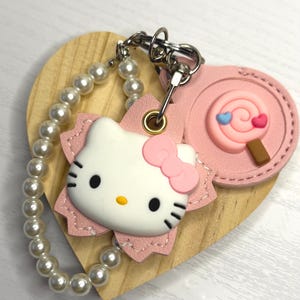 May include: A Hello Kitty keyring in pink and white, with a pearl bead wrist strap. The keyring features a pink bow, black eyes, and whiskers. A pink lollipop charm is also attached. The keyring is attached to a wooden heart.