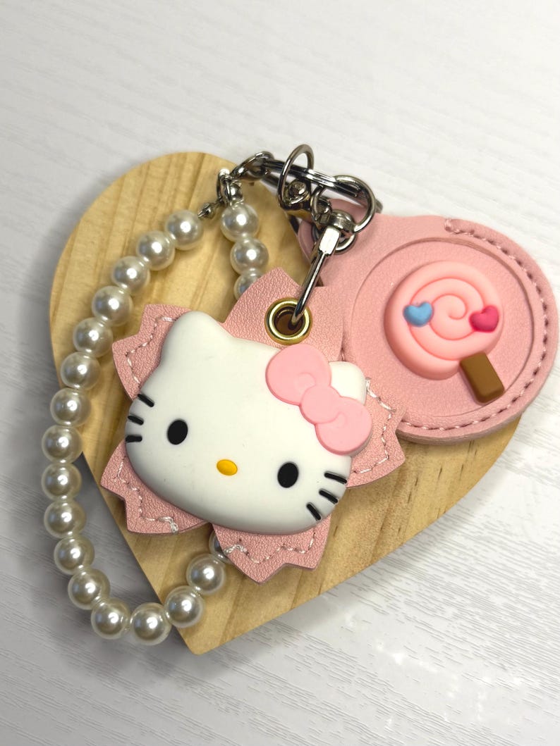 May include: A Hello Kitty keyring in pink and white, with a pearl bead wrist strap. The keyring features a pink bow, black eyes, and whiskers. A pink lollipop charm is also attached. The keyring is attached to a wooden heart.