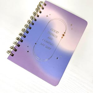 May include: A spiral-bound notebook with a purple and blue gradient cover. The cover has gold foil text reading "GOOD THINGS ARE COMING MY WAY" within a gold frame with star accents. The notebook's binding is gold.