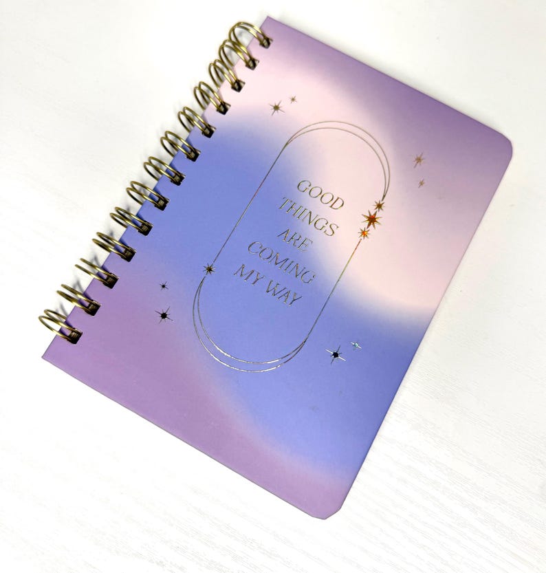 May include: A spiral-bound notebook with a purple and blue gradient cover. The cover has gold foil text reading "GOOD THINGS ARE COMING MY WAY" within a gold frame with star accents. The notebook's binding is gold.