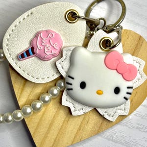 May include: A handmade keychain with a wooden heart, a white leather tag with a pink cotton candy charm, and a Hello Kitty charm with a pink bow. It has a pearl bead chain and a metal clasp.