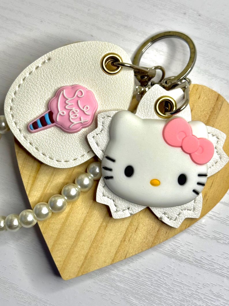 May include: A handmade keychain with a wooden heart, a white leather tag with a pink cotton candy charm, and a Hello Kitty charm with a pink bow. It has a pearl bead chain and a metal clasp.
