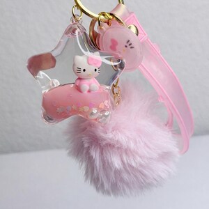 May include: A pink keychain with a star-shaped charm containing a Hello Kitty figure. The keychain includes a fluffy pink pom-pom, a pink strap, and a gold-coloured keyring.