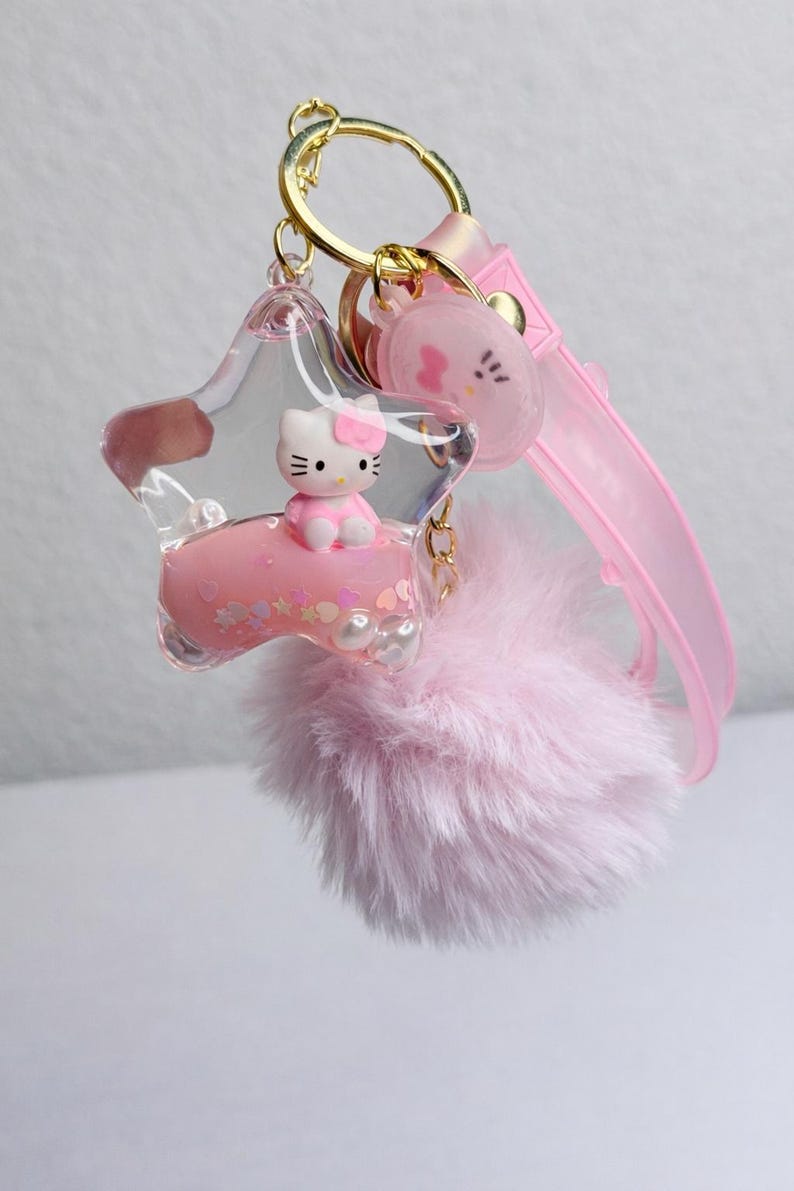 May include: A pink keychain with a star-shaped charm containing a Hello Kitty figure. The keychain includes a fluffy pink pom-pom, a pink strap, and a gold-coloured keyring.