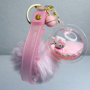 May include: A pink keychain with a Hello Kitty figure in a clear orb filled with pink beads and glitter. The keychain has a pink strap with "MEOW" text and a fluffy pink pom-pom.