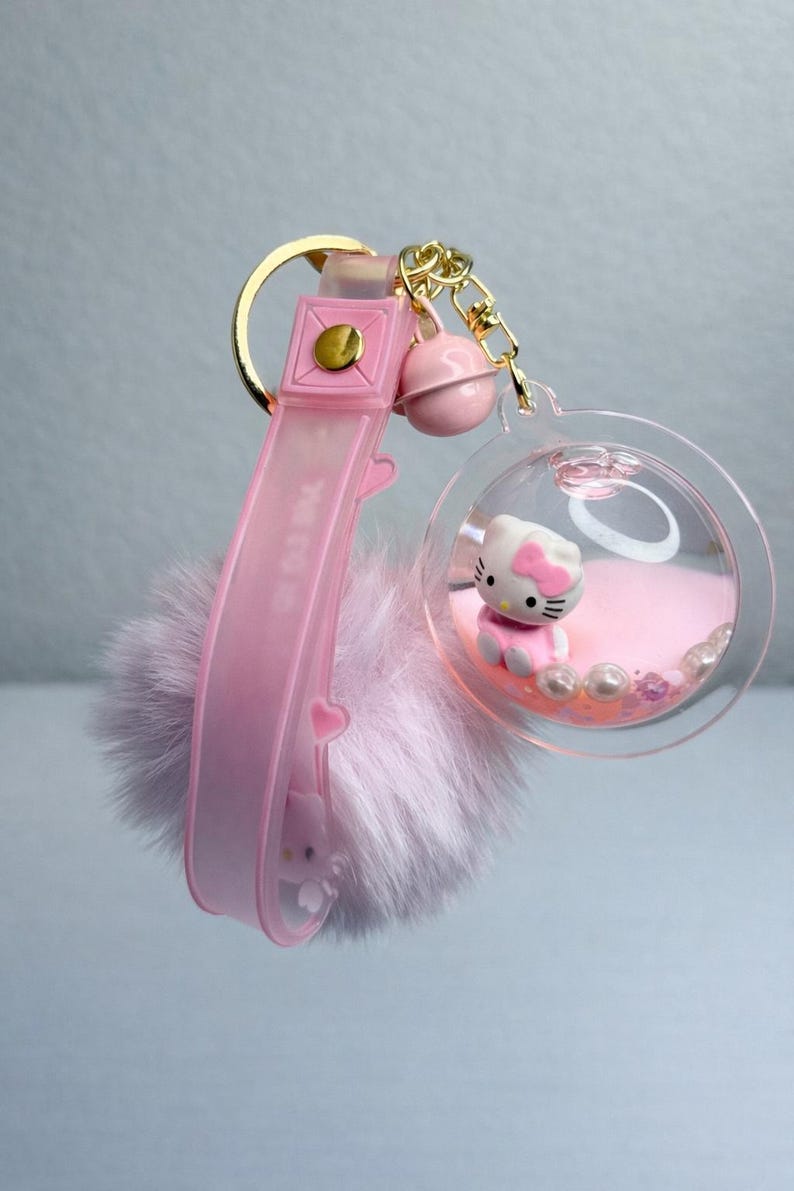 May include: A pink keychain with a Hello Kitty figure in a clear orb filled with pink beads and glitter. The keychain has a pink strap with "MEOW" text and a fluffy pink pom-pom.
